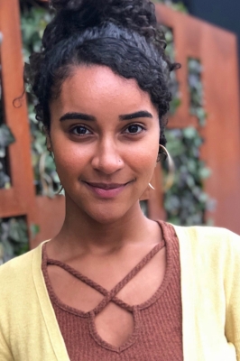 Image of a smiling young woman of color with curly black hair in a top bun, a brown shirt and yellow cardigan