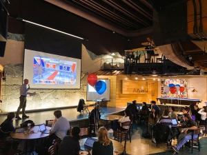 Photo of students sitting at tables at an Election Watch party, watching the results of the election. A man stands on stage and speaks into a microphone.