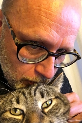 A close up photo of a man leaning forward to put his face near his grey tabby cat. The man is wearing dark-rimmed glasses and is petting the cat's face.