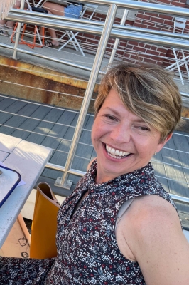 An image of a woman with short, blond hair is smiling warmly into the camera. She is wearing a summer dress that is brown with small blue flowers. She is sitting at a cafe table outside.