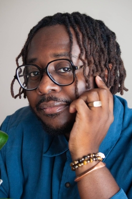 A black man with chin-length black hair, a beard, large black glasses, a blue button-down shirt, and gold and beaded bracelets, leans forward to rest his chin in his hand. He is looking into the camera.