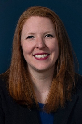 A white woman with red hair, a blue shirt, and a black blazer smiles into the camera.