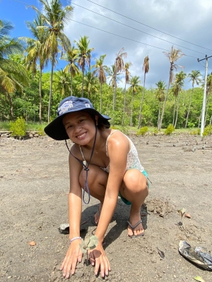 A young woman crouching down on the beach planting a tree. She is wearing shorts and a tank top and bucket hat. She is smiling into the camera.