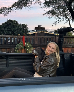 Anna smiles in the back of a pickup truck bed hugging a black dog under a tree. She has blonde hair and is wearing a leather jacket.