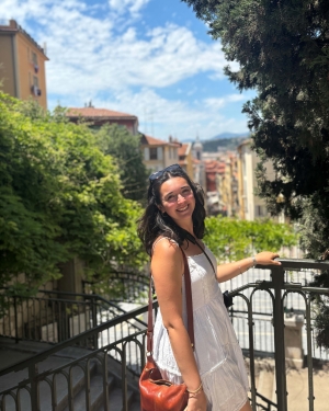 Joly stands in a white dress overlooking a viewpoint with trees and buildings in the background. Her hair is shoulder-length and brown and her hand is on the railing beside her.