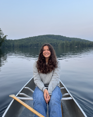 Paige sits with long brown hair and a gray shirt in a canoe on a lake with a cloudy sky behind her