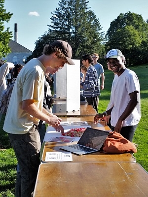 Two people stand outside on either side of a table, one looking at the camera and smiling, the other looking down at a piece of paper. Other people are visible in the background, as well as grass and trees.