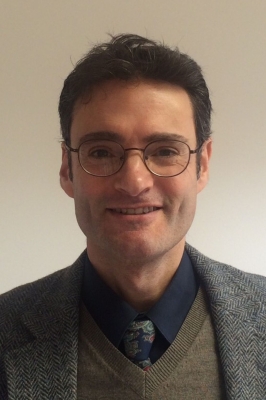 A man with short, brown hair, dimples, and wire-framed glasses smiles into the camera. He is wearing a navy blue button-down, a blue-patterned tie, an olive v-neck sweater, and a grey wool suit jacket.