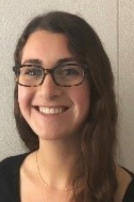 A young woman with long brown hair, a black v-neck top and tortoise shell glasses smiles into the camera.