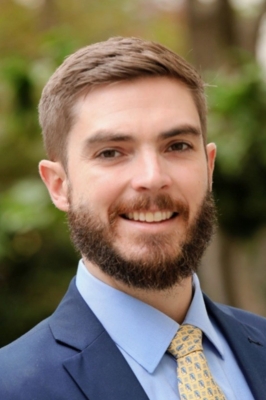 A man with short brown hair and a brown beard, wearing a light blue button-down shirt, yellow tie, and navy blue blazer smiles into the camera.