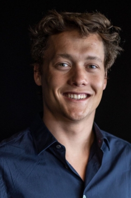 A young man with a navy blue button-down shirt and short, brown hair, smiles into the camera.