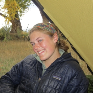 A person with blonde hair tied in a multicolored bandana wearing a black jacket is smiling.