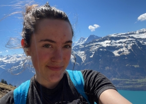 A selfie of a person wearing a blue back pack standing in front of some mountains.