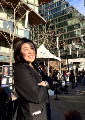 A girl with black hair in a black suit jacket is standing with arms crossed and smiling.
