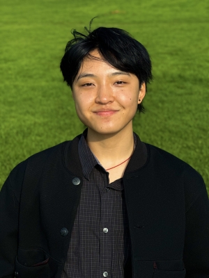 A person with black hair, a black shirt and a black coat standing in front of a green grass background, smiling.