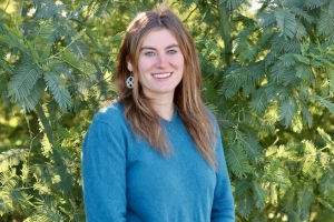 A person with long brown hair and a blue top is standing in front of a leafy green background, smiling.