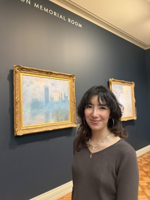 A person with dark hair in a grey shirt is smiling while standing in front of an art piece.