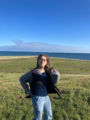 A person in keans and a blue shirt are standing in front of a grassy plain with a body of water in the background.