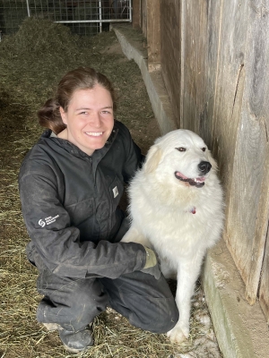 A person in full black clothing is sitting near a white dog.