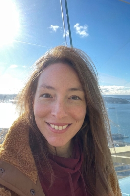 A woman with long, straight, brown hair, stands outside in front of water, with a brown fleece jacket on and brown scarf. She is smiling into the camera.