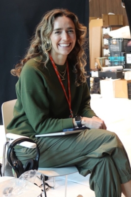 A young woman with long, wavy brown hair, wearing army green pants and a green sweater, sits in an office chair with her leggs crossed and smiling into the camera.