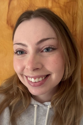 A young woman with long dark blonde hair and a grey hooded sweatshirt stands in front of a wood wall and smiles into the camera.