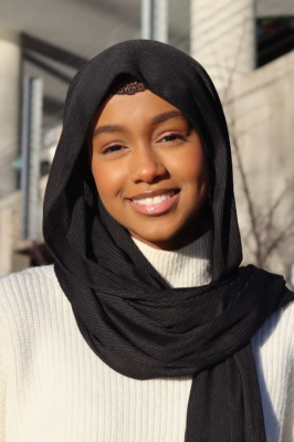 A young woman with a black hijab and a white knit turtleneck. She is smiling into the camera.