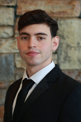 A young man with short black hair, a white button-down shirt and black tie, wears a black suitcoat and looks into the camera. He is standing in front of a stone wall.