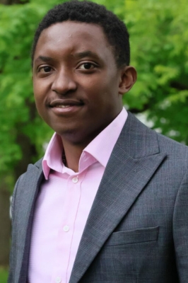 A young black man with a pink button-down shirt and grey suit coat stands outside in front of a tree. He is smiling into the camera.