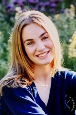 A young white woman is seated outside in front of a flower garden. SHe is wearing a navy blue v-neck shirt and has long dirty-blond hair. She is smiling into the camera.
