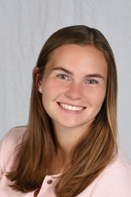 A young white woman with a light pink button-down shirt and long dark blonde hair smiles into the camera.