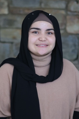 A young woman with a black hijab and tan turtleneck smiles into the camera. She is standing in front of a stone wall.