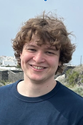 A young man with blond curly hair and a navy blue t-shirt stands outside and smiles into the camera.