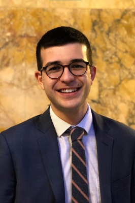 A young man with short, black hair and a navy blue suit coat, with a white button-down shirt and blue and yellow tie, smiles into the camera.