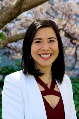 A young woman poses in front of a tree in full blossom. She has shoulder-length black hair, a white blazer and a dark red dress. She is smiling into the camera.
