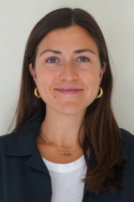 A woman with long, straight, brown hair, gold hoop earrings, a white t-shirt and a blue blazer smiles into the camera