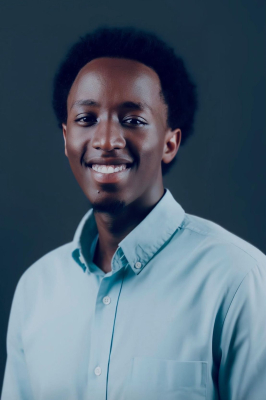 A young man with black hair and a blue button-down shirt smiles warmly into the camera.