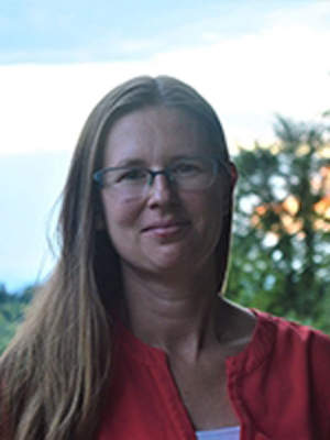 Amanda Crocker, long, straight, brown hair, wearing glasses and red shirt. There is sky and trees behind. 
