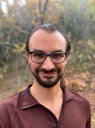 Phil Chodrow, short dark hair, glasses, mustache and beard burgundy collared shirt with trees in background