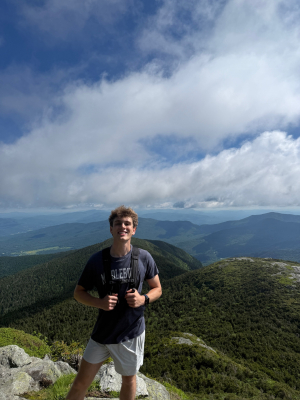 Young man with dirty blonde hair and a Middlebury shirt stands on a mountain smiling. 