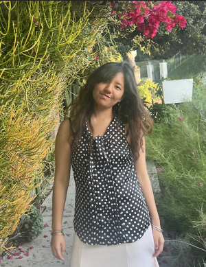 Young woman with dark brown hair and a polka dot blouse smiles with a colorful garden behind her. 