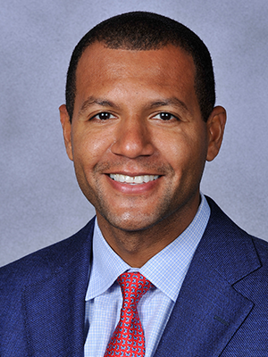 Koby Altman headshot, wearing a dark blue suit, light blue shirt, red tie