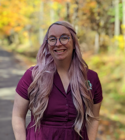 Young woman with glasses and long curled purple hair standing in front of yellow, green, and orange trees