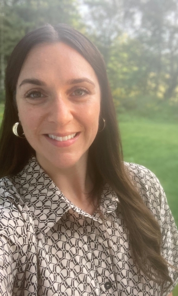 A smiliing person with Dark hair and hoop earrings in front of trees and grass