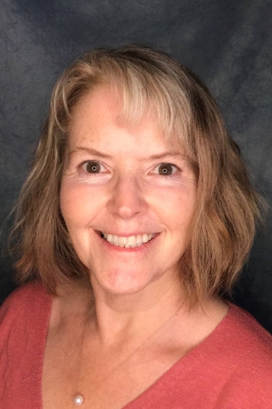 Photo of a woman with shoulder-length, wavy blond hair and bangs. She is wearing a peach v-neck sweater and gold pearl necklace. She is smiling into the camera.