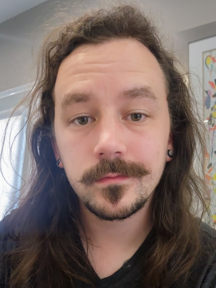 Headshot of Mathew Muston - a man with long brown hair and facial hair, wearing a black shirt