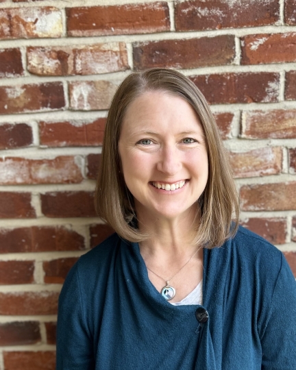 Sarah Messner, wearing a blue shirt, smiles and poses for a portrait in front of a red and white brick wall.