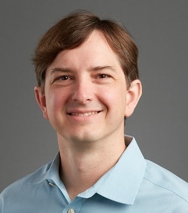 A smiling man with short brown har and a blue collared shirt.