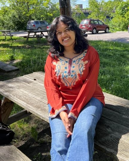Priya smiling outside sitting on a picnic table with an orange shirt and grass in the background
