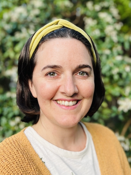 A person in a yellow shirt and yellow headband smiles at the camera, in front of some greenery.